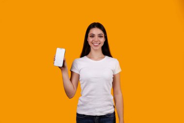 Uzun saçlı, beyaz tişörtlü güzel esmer bir kadın izole edilmiş sarı arka planda telefonla poz veriyor. Boş cep telefonunu gösteren kadın modelin portresi. Kapat, boşluğu kopyala.