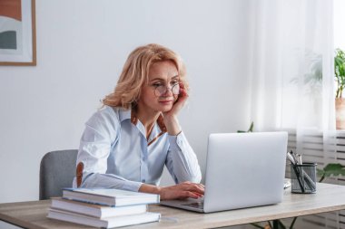 İş yerinde oturan mavi bluz ve gözlüklü yetişkin sarışın kadın portresi. Ofiste dizüstü bilgisayarla çalışan zeki bir iş kadını. Arkaplan, kopyala boşluk, kapat.
