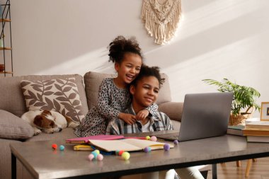 Farklı iki güzel siyah kız ve tapılası tel saçlı Jack Russel Terrier köpekleri evde dizüstü bilgisayar kullanıyorlar. Sert kaplamalı sevimli kız kardeşler eğleniyor. Arkaplan, kapat, boşluğu kopyala.