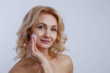 Tamamen temiz ciltli yetişkin bir kadının portresi pamukla losyon sürüyor. Dalgalı sarı saçlı, gülümseyen ve günlük cilt bakımını yapan bir kadın. Kapat, arkaplanı kopyala.