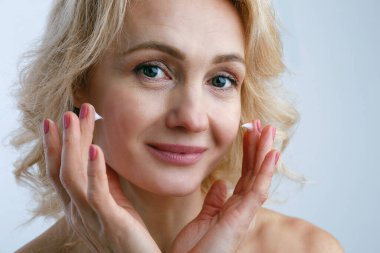 Nemlendirici krem sürerek cildini temizleyen yetişkin bir kadının portresi. Orta yaşlı, dalgalı sarı saçlı, gülümseyen, günlük cilt bakımını yapan bir kadın. Kapat, arkaplanı kopyala.