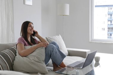 Genç esmer kadının portresi oturma odasındaki koltukta oturuyor, dizüstü bilgisayarda çalışıyor, telefonla konuşuyor ve gülümsüyor. Neşeli kadın, eğlenceli bir telefon görüşmesi yapıyor. Arkaplan, kopyala boşluk, kapat.