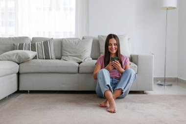 Genç esmer kadın portresi, anne kotu giymiş, yere oturmuş kanepeye yaslanmış, mesajlaşıyor ve gülümsüyor. Neşeli kadın, eğlenceli bir telefon görüşmesi yapıyor. Arkaplan, kopyala boşluk, kapat.