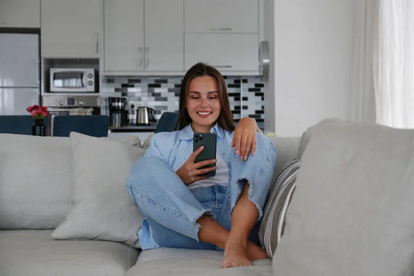穿着妈妈牛仔裤躺在沙发上发短信微笑的年轻黑发女人的画像 快乐的女性有一个有趣的视频电话 复制空间 — 图库照片