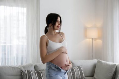 Hamileliğin ikinci üç ayında genç ve güzel bir kadın. Hamile kadın destekleyici, dikişsiz annelik sutyeni giyiyor ve kolları göbeğinde olan fermuarsız anne pantolonu giyiyor. Bir çocuk bekliyorum. Arkaplan, boşluğu kopyala