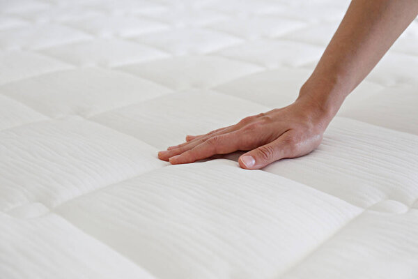 Cropped shot of young woman's hand testing white orthopedic matress on firmness. Female pressing hypoallergenic foam mattress surface to check its softness. Close up, copy space, top view, background.