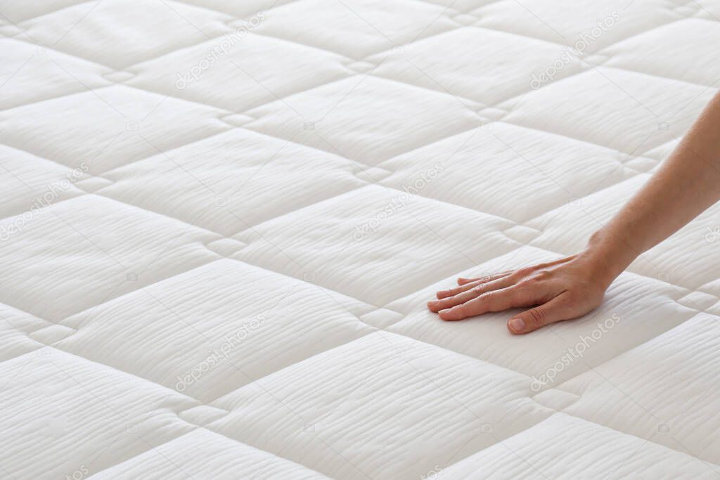 Cropped shot of young woman's hand testing white orthopedic matress on firmness. Female pressing hypoallergenic foam mattress surface to check its softness. Close up, copy space, top view, background.
