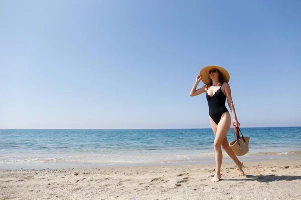 若い美しい女性はビーチバッグでポーズを1ピース水着 広いつばのわら帽子を着用 美しい晴れた日に砂浜で女性ファッションモデル 地中海の海の背景 スペースのコピー — ストック写真