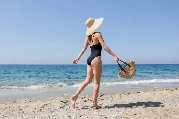 若い美しい女性はビーチバッグでポーズを1ピース水着 広いつばのわら帽子を着用 美しい晴れた日に砂浜で女性ファッションモデル 地中海の海の背景 スペースのコピー — ストック写真