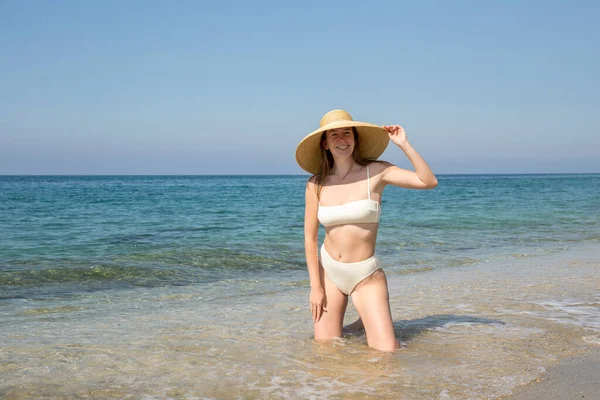 Güzel, güneşli bir günde kumsalda poz veren, yüksek bel altı ve geniş kenarlı hasır şapkalı, sağlıklı vücutlu, bikinili beyaz bir kadın. Akdeniz arka planı. Metin için alanı kopyala