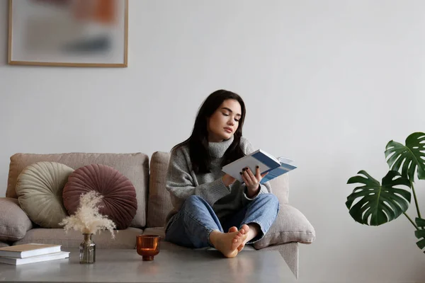 年轻漂亮的女人穿着灰色的高领毛衣在家里看书 穿着毛衣的布鲁内特女坐在她高贵公寓里的米色纺织品沙发上 复制空间 — 图库照片