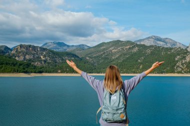 Kapüşonlu ve sırt çantalı bir kadın gezgin dağ gölü manzarasının keyfini çıkarıyor. Genç bir kadın doğada yalnız kalmaktan ruhani bir mutluluk yaşıyor. Arkaplan, kopyalama alanı, arka görünüm.