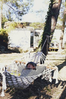 Hippi kıyafeti giyen ve şapkası olan rahat genç bir adam güzel güneşli bir günde hamakta çıplak ayakla tembelce yalnız kalmanın tadını çıkarıyor. Yavaş yaşam konsepti. Kapat, arkaplanı kopyala.