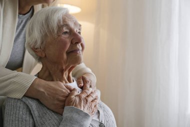 Unrecognizable female expressing care towards an elderly lady, hugging her from behind holding hands. Two adult women of different age. Family values concept. lose up, copy space, background.