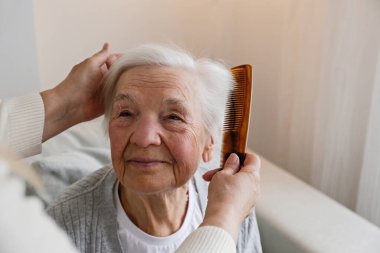 Unrecognizable female expressing care towards an elderly lady, brushing her hair with a comb. Granddaughter helping granny with a haircut. Family values concept. lose up, copy space, background.