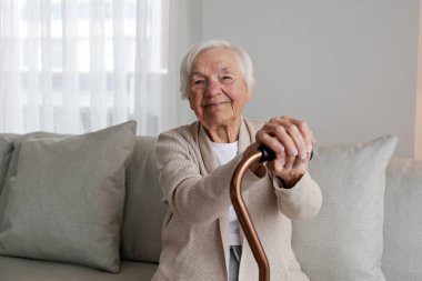 Portrait of a senior female sitting on the couch at home, leaning on a cane. Elderly woman holding the wooden handle of a metal walking cane. Close up, copy space for text, background.