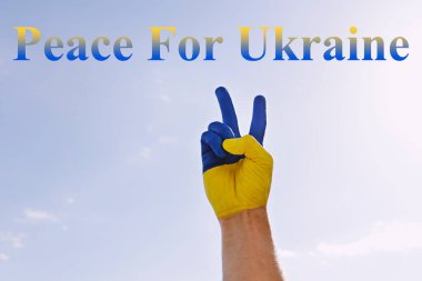 Mavi gökyüzü arka planında yazan Ukrayna için barış. Ukrayna ulusal bayrağına boyanmış bir adamın eli Ukrayna 'ya destek ve dayanışma göstergesi olarak barış jesti sergiliyor. Boşluğu kopyala.