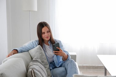 Anne kotu giyen esmer bir kadının portresi kanepede oturmuş mesaj atıyor ve gülümsüyor. Neşeli kadın evde videolu telefon görüşmesi yapıyor. Arkaplan, kopyala boşluk, kapat.