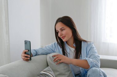 Anne kotu giyen esmer bir kadının portresi kanepede oturmuş mesaj atıyor ve gülümsüyor. Neşeli kadın evde videolu telefon görüşmesi yapıyor. Arkaplan, kopyala boşluk, kapat.