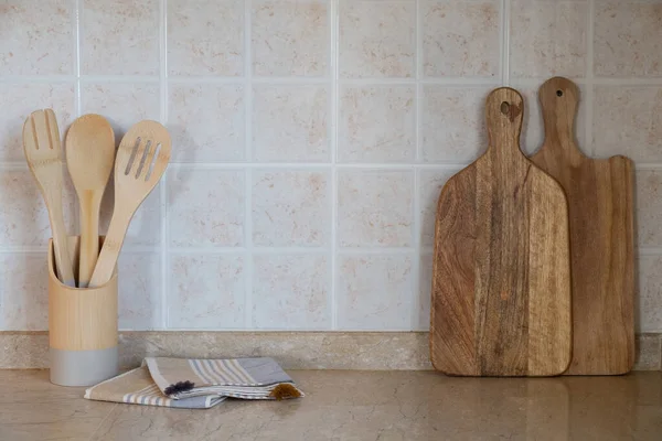 Different kitchen utensil made of wood in a holder, cutting board and a blanket on marble tabletop. Copy space for text, close up, wall with beige facing tile background.