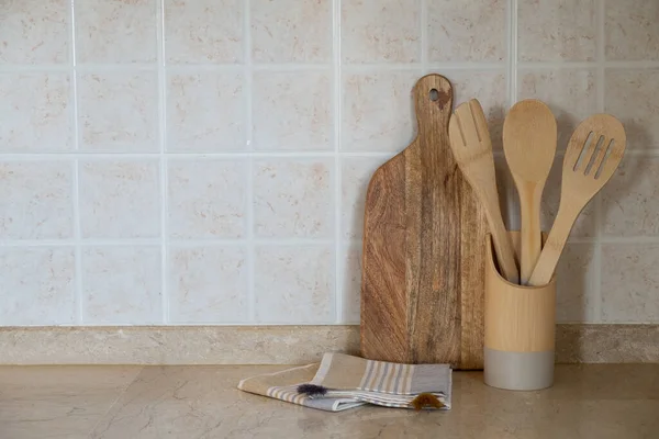 Different kitchen utensil made of wood in a holder, cutting board and a blanket on marble tabletop. Copy space for text, close up, wall with beige facing tile background.