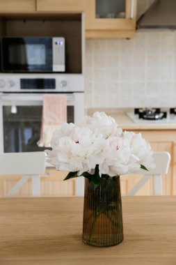 Mutfak masasındaki cam vazoda güzel şakayık çiçekleri. Küçük bej fayanslı ve ahşap dolaplı modern mutfakta yemyeşil bir buket yakın çekim. Kapat, kopyala boşluk, arkaplan.