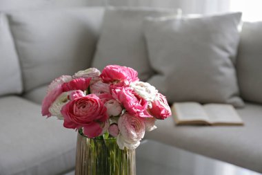 Ön planda vazoda iki renkli ranunculus çiçekleri ve arka planda gri tekstil kanepe olan cam sehpanın yakın çekimi. Uzayı kopyala, kapat, doğal ışık.