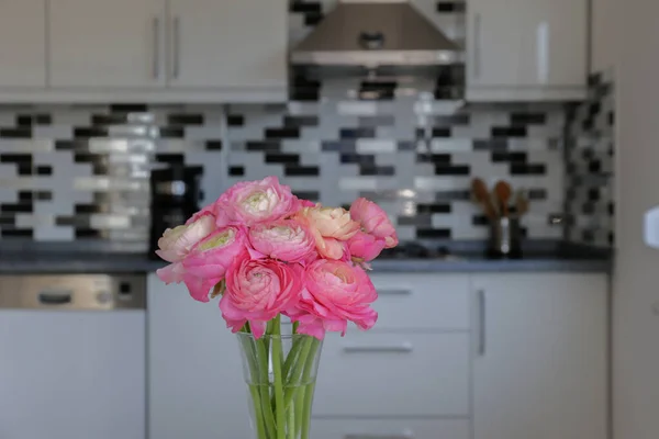 Bouquet of pink ranunculus flowers in glass vase on the kitchen counter. Stylish modern kitchen with small mosaic tile and modern white cupboard. Close up, copy space, background.
