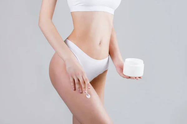 Studio shot of fit woman posing over isolated white background, applying moisturizing body lotion to her leg. Female in white underwear holding a jar with a skincare product. Copy space, close up.