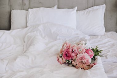 Beyaz çarşaflı boş yatağın üzerinde harika bir buket şakayık ve ranunculus çiçeği yakın çekim. Romantik bir sabah sürprizi. Metin, arkaplan, üst görünüm, doğal ışık için alanı kopyala.
