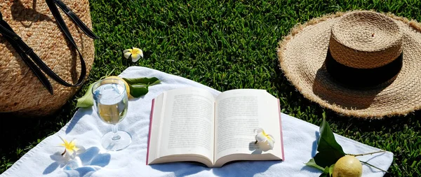 Lunch break in the park. Picnic blanket with an open book, lemons, beach bag and broad brim straw hat on a juicy green freshly freshly mowed lawn. Close up, copy space, top view, background.