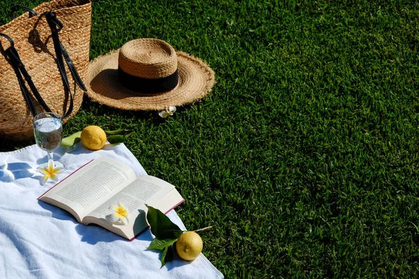 Lunch break in the park. Picnic blanket with an open book, lemons, beach bag and broad brim straw hat on a juicy green freshly freshly mowed lawn. Close up, copy space, top view, background.