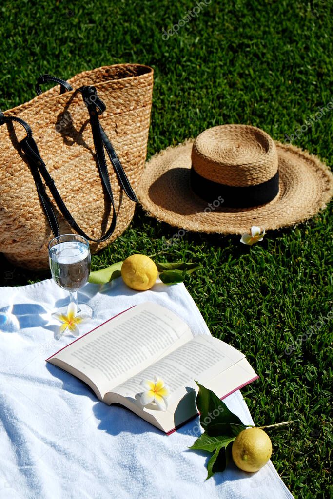 Lunch break in the park. Picnic blanket with an open book, lemons, beach bag and broad brim straw hat on a juicy green freshly freshly mowed lawn. Close up, copy space, top view, background.