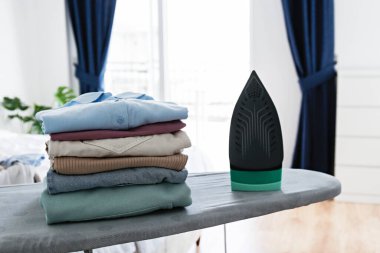 Evde temiz, yeni yıkanmış giysiler ve elektrikli ütü yığını var. Ütü masasında bir yığın gömlek, beyaz perdeli uzun bir pencere. Boşluğu kopyala, kapat, üst görünüm, arkaplan