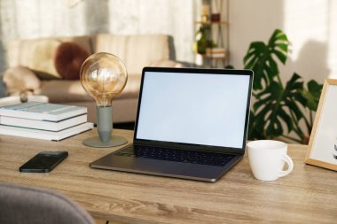 Blank screen laptop, cell phone and a cup on wooden table. Cozy lofty office with house plants full of natural sunlight. Close up, copy space, background.