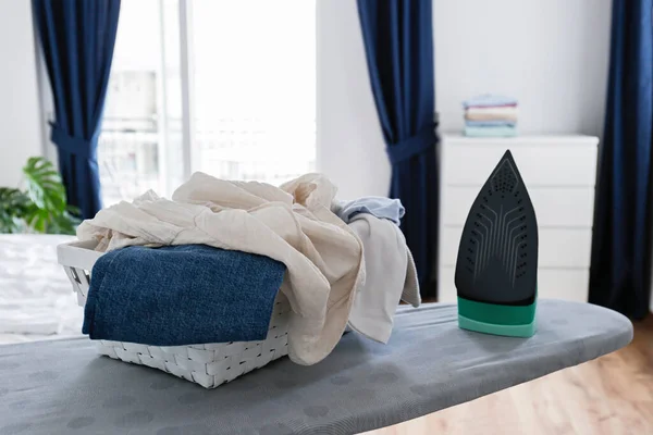 Neat stack of clean freshly laundered clothes and electric iron on pressboard in living room. Pile of t-shirts and sweaters on ironing board, home interior background. Copy space, close up, top.