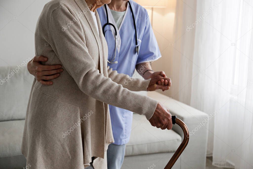 Senior woman with a cane getting assistance in elderly care facility. Hospital nurse taking care of mature female with walking stick in nursing home. Background, close up on hands with wrinkled skin.