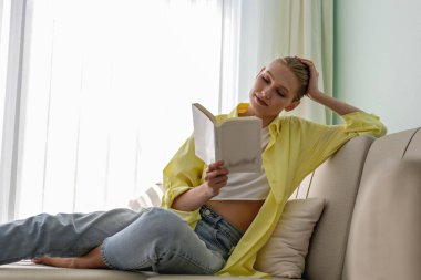 Sarı gömlekli genç bir kadın evde kitap okuyor. Esmer kadın, yüksek dairesinde tekstil kanepesinde yatıyor ve ders çalışıyor. Arkaplan, kopyala boşluk, kapat.