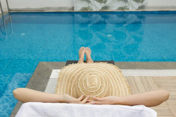 Young woman with fit body wearing purple bikini swimsuit and covering her face with broad brim straw hat as protection from the sun. Sunbathing by the swimming pool. Copy space.