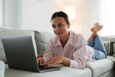 Close up shot of young woman working remotely from home on laptop, lying on the blue couch in living room. Female freelancer with hands on keyboard. Copy space, background