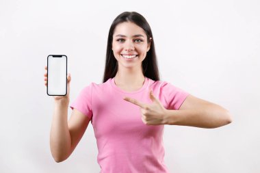 Young beautiful brunette woman with long hair wearing pink t-shirt, pointing at a phone over isolated background. Portrait of female model showing blank screen cellphone. Close up, copy space