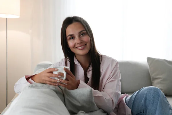 Portrait of young beautiful woman wearing a pink blouse, sitting on the couch holding a cup of coffee. Brown eyed female with smiling facial expression. Copy space, background, close up