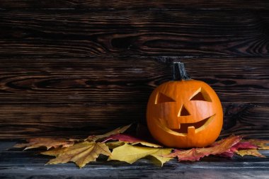 Big orange pumpkin carved into Jack o Lantern and dry maple leaves laying on wooden table. Background, copy space, close up, top view. All hallows eve Halloween party decoration concept.