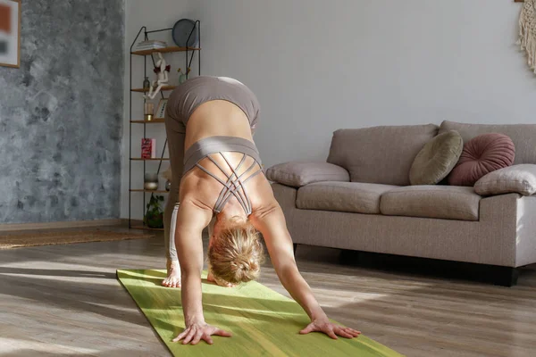 Sporty adult woman practicing hatha yoga at home. Fit middle aged yogini, doing the downward facing dog pose. Interior background, copy space, close up.