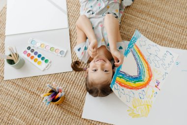 Çocuk yerde yatıyor ve çizimini gökkuşağıyla yapıyor.
