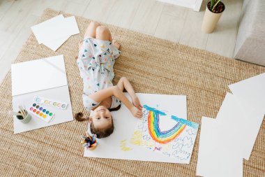 Çocuk yerde yatıyor ve çizimini gökkuşağıyla yapıyor.