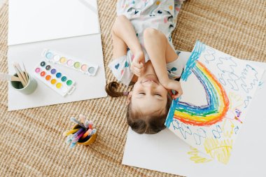 Çocuk yerde yatıyor ve çizimini gökkuşağıyla yapıyor.