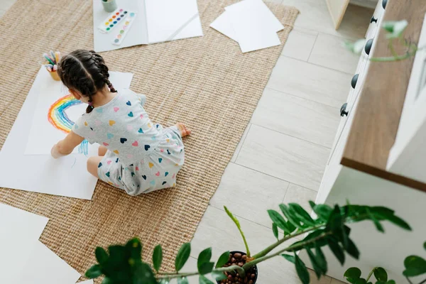 Çocuk yerde yatıyor ve çizimini gökkuşağıyla yapıyor.
