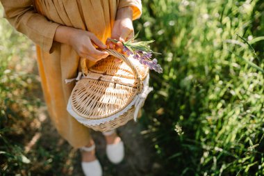 Summer vibes. The feeling of summer. Unrecognizable little girl wearing linen mustard dress walking on the path with basket and lupine flowers