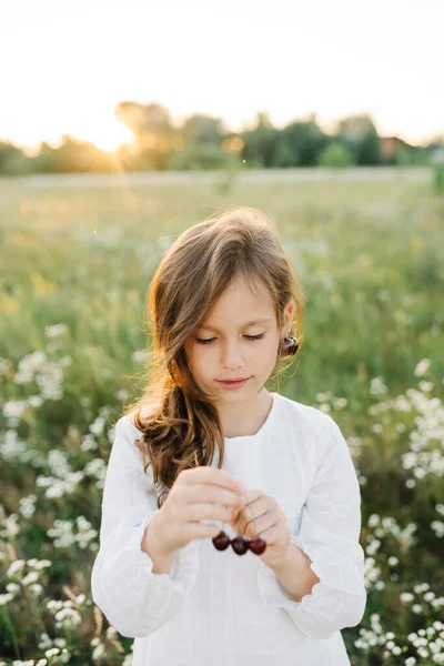 Summer vibes. The feeling of summer. Portrait of cute little girl wearing white blouse in the fields having cherries earrings